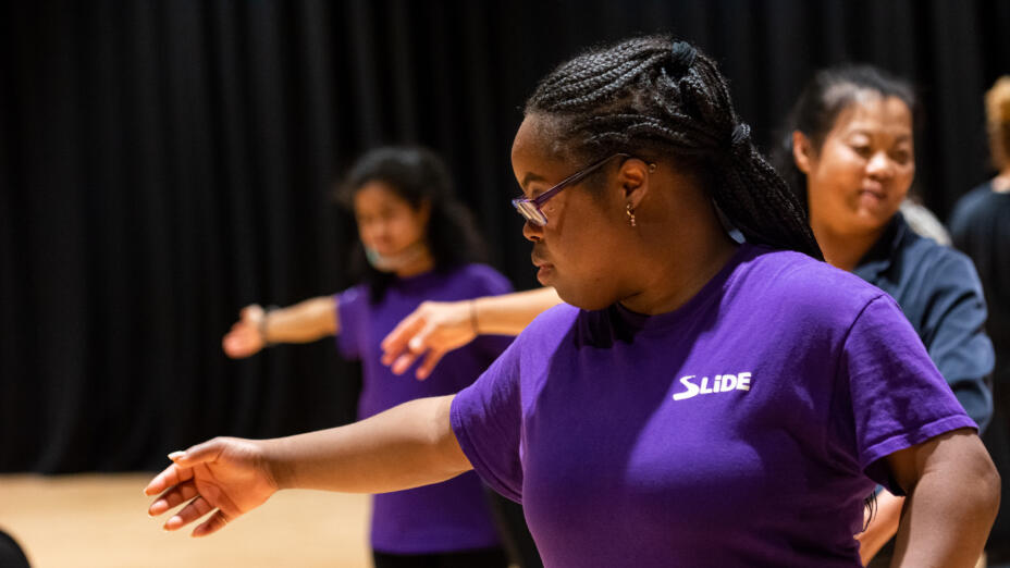 Young people are rehearing a dance in a black studio room. The girl at the front is wearing a purple t-shirt and is posing with her right arm stretched out in front of her, with her left arm more relaxed by her side