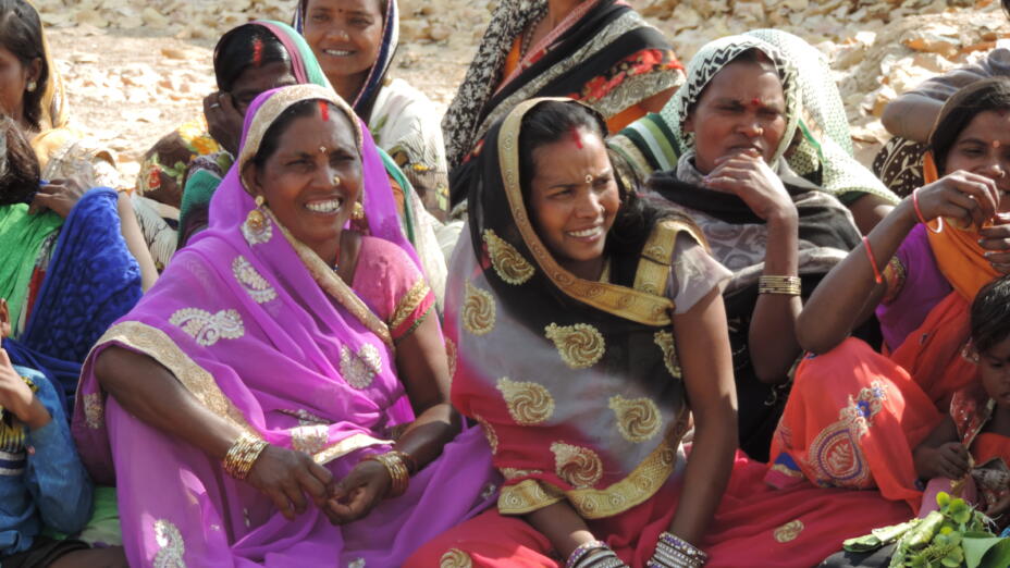 Group of women wearing colourful saris embroidered with gold. They are sitting on the ground and are smiling and listening.