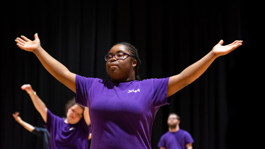A group of four young people are in a dance studio with wood flooring and black walls. A young woman is in the front, in a purple t-shirt with her arms raised up to her sides