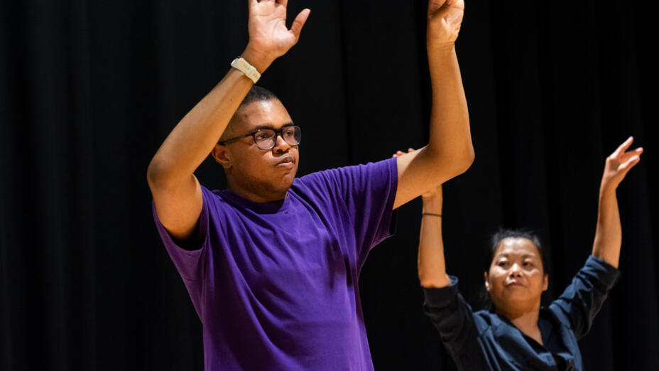 Two people are in a dance studio with wood flooring and black walls. One young man is in the front, with arms raised towards the ceiling
