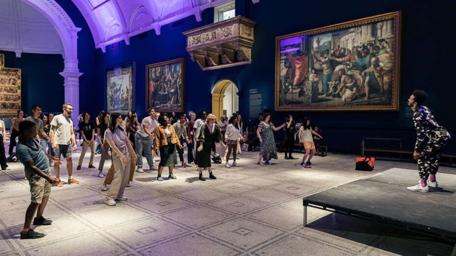 A class of around 30 adults are standing in a dark blue room in the V&A, with paintings in the background. A young man leading the class is at the front on a small stage, teaching a dance