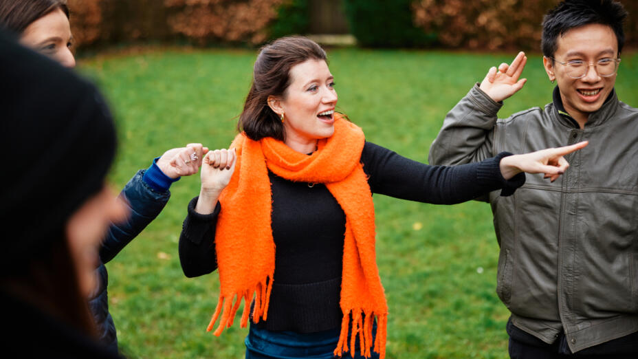 A group is outdoors, surrounded by trees and grass. They are standing in a circle, smiling and playing a game where they are touching each other's hands