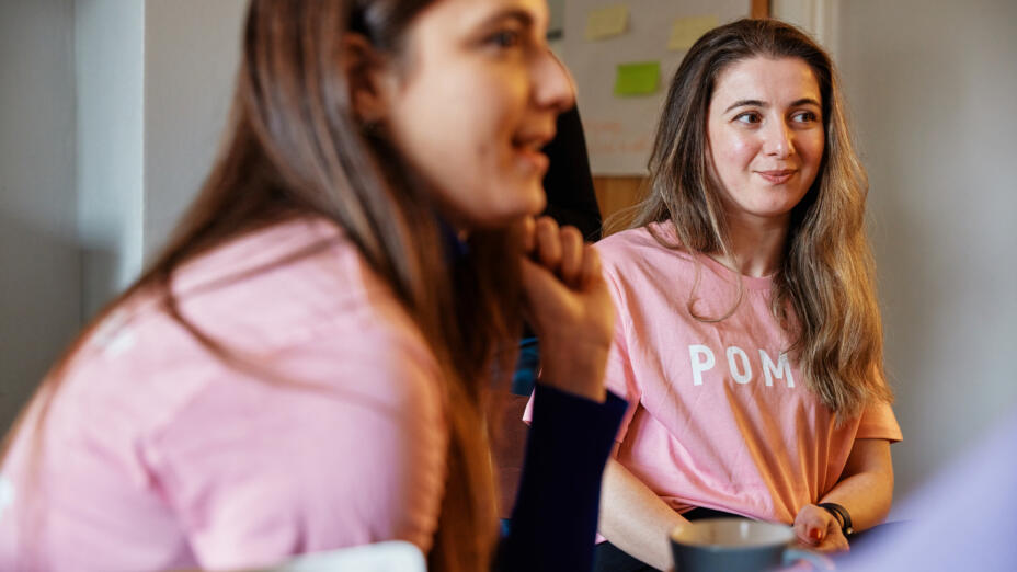 Two young women are in conversation with a group, wearing pale pink t-shirts that say POMOC