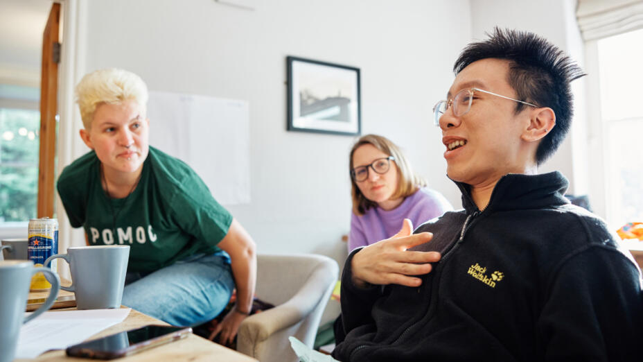 Three people are sitting in a living room, with mugs and papers on a table in front of them. A young man is speaking and smiling with two people listening