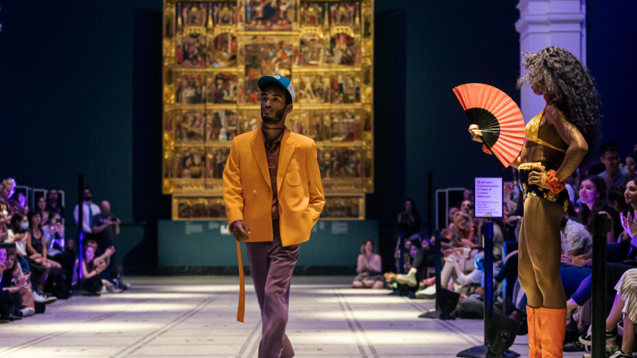 A young man is walking a runway, with a seated audience either side. He is wearing a bright yellow jacket with a long belt, purple trousers and baseball cap. A young woman is standing to the side with bright orange boots, a red fan and a yellow and black outfit