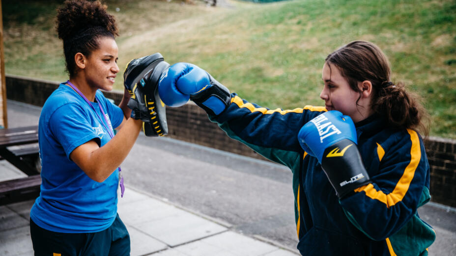 Boxing coach wearing a blue t-shirt holds a training session outside with a young girl wearing blue boxing gloves.