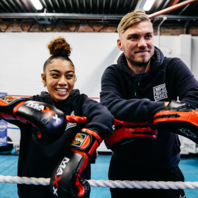 Young man and woman in a boxing ring, leaning on the ropes and smiling off-camera.