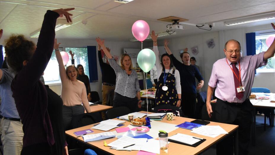 A group of teachers dance around a table joyously with their arms in the air. There are balloons, a cake and papers on the table.