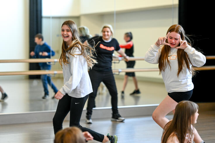 Two young girls are mid-dance in a dance studio. A teacher is behind them. One girl is smiling, mid-step, and the other is holding their hands up in a frame pose to their face