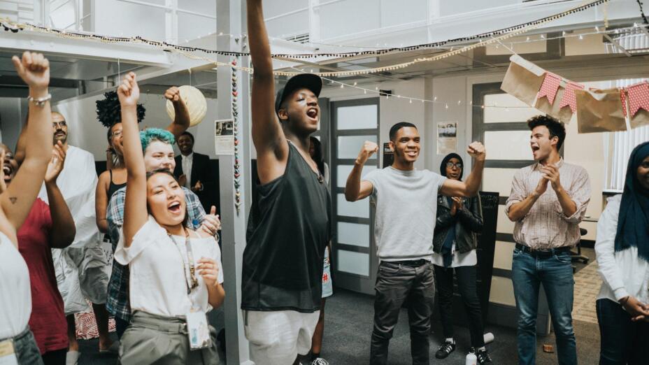 A group of a dozen or so young people are in a room, all cheering. Some have their arms stretched out above their heads while others are clapping