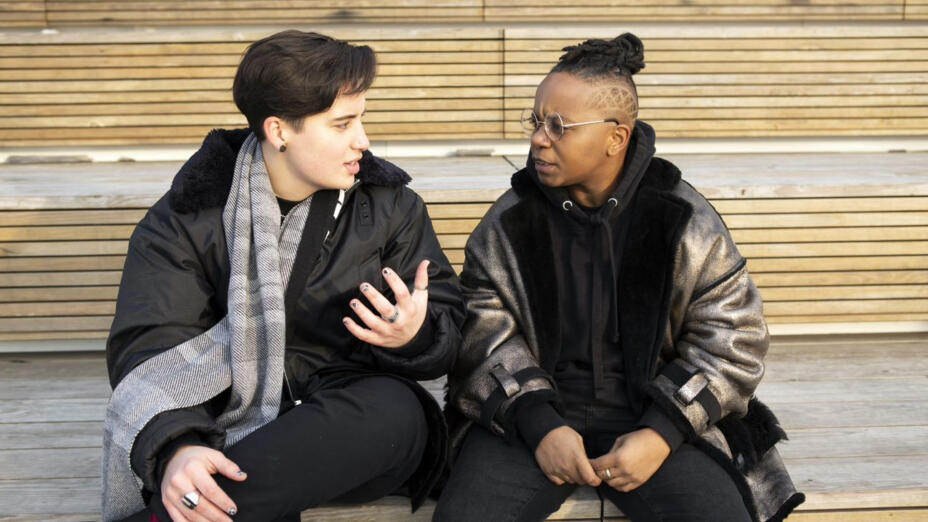 Two people are sat on a wooden structure outside, in conversation