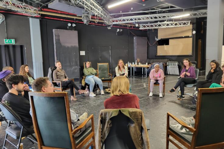 A group of teachers sit on chairs in a circle in a rehearsal hall with a concrete floor and dark grey walls.
