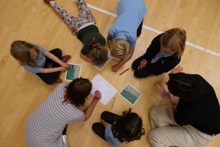 Overhead photo of 5 children and 2 teachers sitting on the floor with pencils looking at photographs as stimulus for a creative writing session.