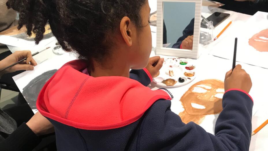 A young girl is in a painting class, holding a mirror and starting to paint a self-portrait