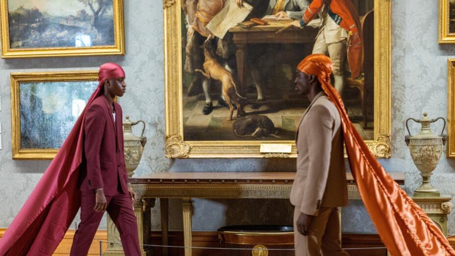 Two young black men are walking towards each other. In the background, there are ornate 18th and 19th century paintings in gold frames from the National Museum Wales collection. The young man on the left is dressed in a plum red suit and a similar coloured durag which flows down into floor-length silk. The man on the right is in a tan suit, with a bright orange durag which similarly flows down into floor-length silk