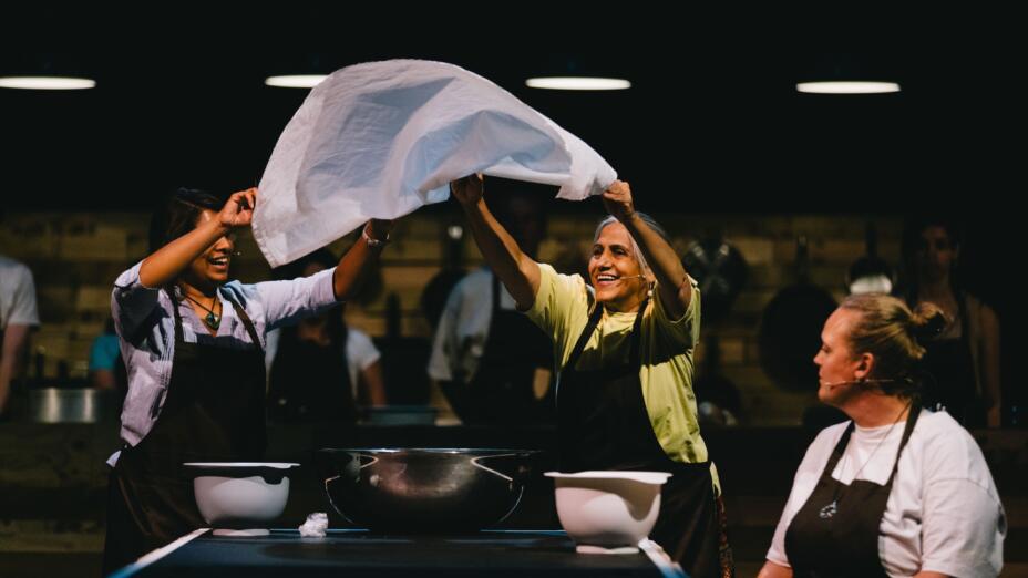 Two smiling women raise a cloth together in the air performing in Born and Bread at Brighton People’s Theatre. Photo: Rosie Powell.