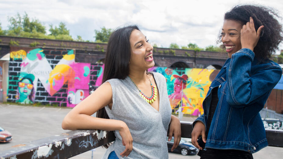 Two young people laughing in front of a wall with graffiti on it.