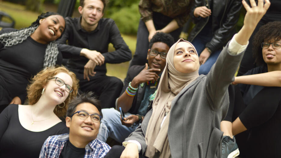 A group of young people sit outside on green grass smiling while a young woman in head scarf takes a group selfie of them.