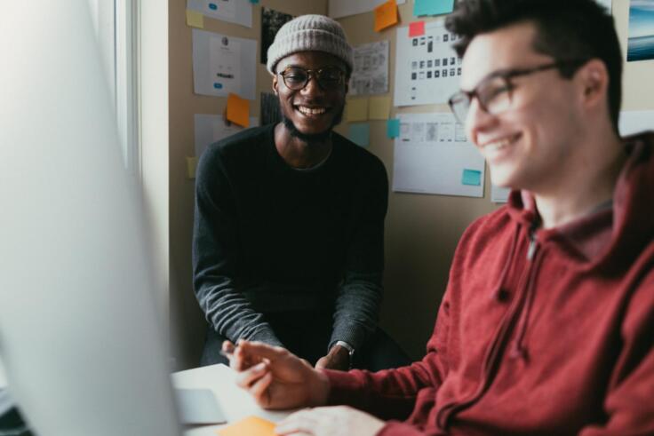Two young men collaborate working at lap top and laughing. One wears a black shirt and a grey beanie and the other wears a red hoodie.