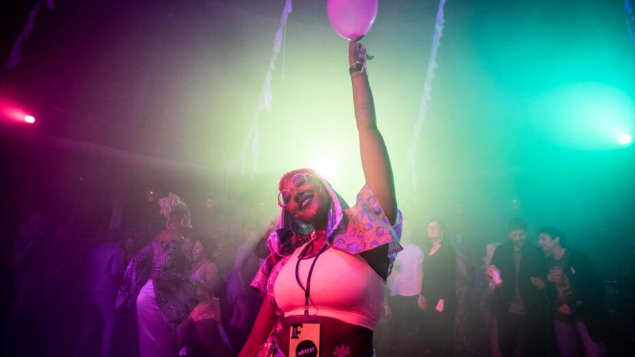 A young person wearing a lanyard that says 'artist' is standing, facing the camera and holding a pink balloon above their head. They are in a nightlife venue with a crowd of people and blue, red and yellow lights.