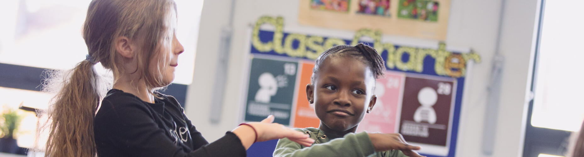 Two young girls are in a classroom, mid-dance. They have their arms crossed out in front of them, one above the other Photo credit: Camilla Adams