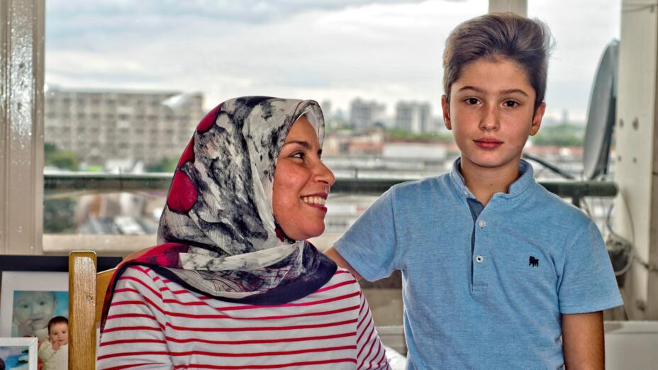 Woman wearing a red stripe shirt and a wearing a red and grey floral hijab. She sits smiling at her son who is standing wearing a light blue button down shirt and faces the camera. They're both sitting in front of a window overlooking a cityscape.