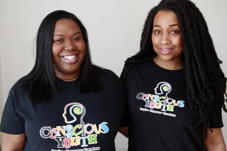 Two Black women smile at the camera with their arms around each other. They are both wearing Conscious Youth t-shirts.