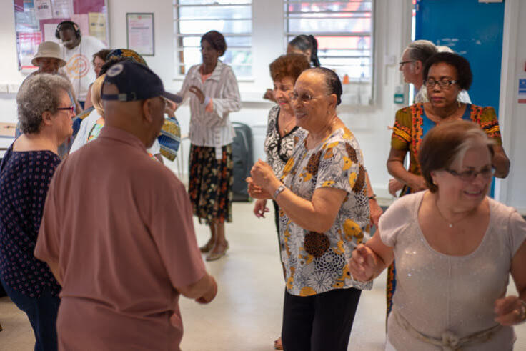 A group of 10 or so older people are standing in a room, talking and smiling, with a DJ playing music in the background