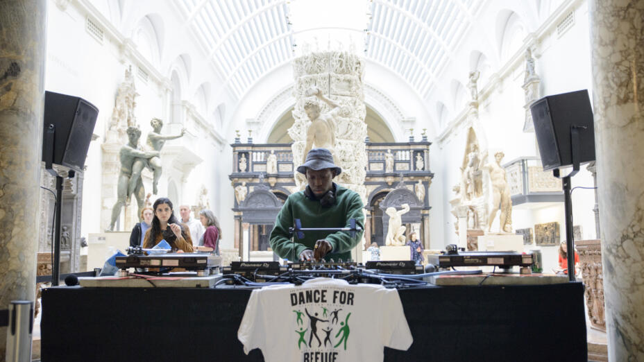 A young man is DJ-ing at the V&A, with a t-shirt on his decks saying 'dance for refuge'. In the background, there are marble pillars and marble statues from the V&A's collection