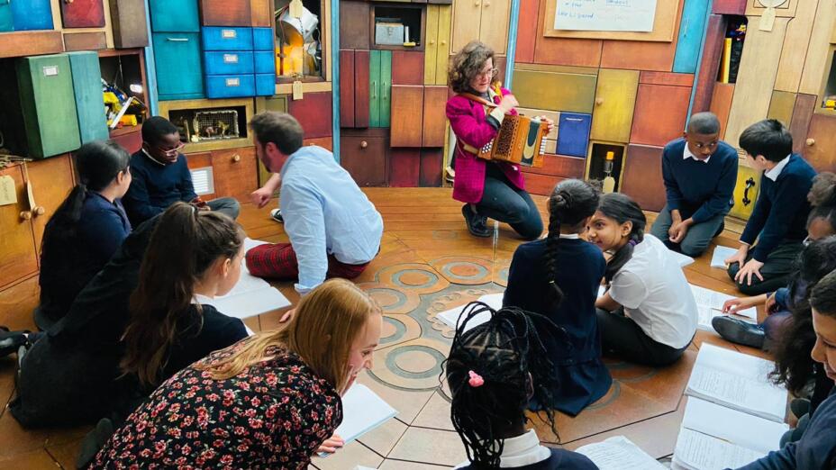 A group of children are sitting in a circular classroom, with a few adults facilitating each small group in the class. A woman at the front is kneeling with an accordion, in front of a whiteboard that has suspense written on it