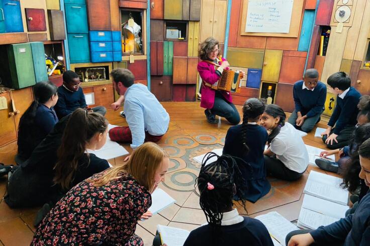 A group of children are sitting in a circular classroom, with a few adults facilitating each small group in the class. A woman at the front is kneeling with an accordion, in front of a whiteboard that has suspense written on it
