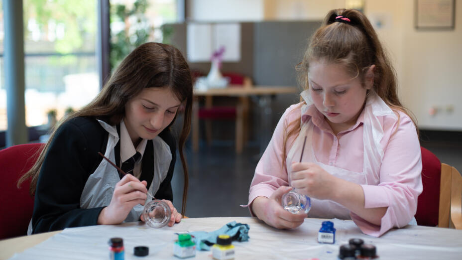 Two young girls are in a classroom, with paints spread out in front of them. They are each painting a glass jar and concentrating on their work