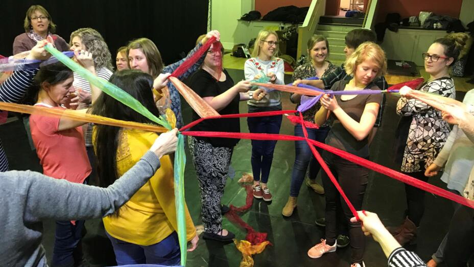A large group of teachers and artists take part in arts-based CPD. The group are holding long pieces of blue, red, green and yellow fabric.