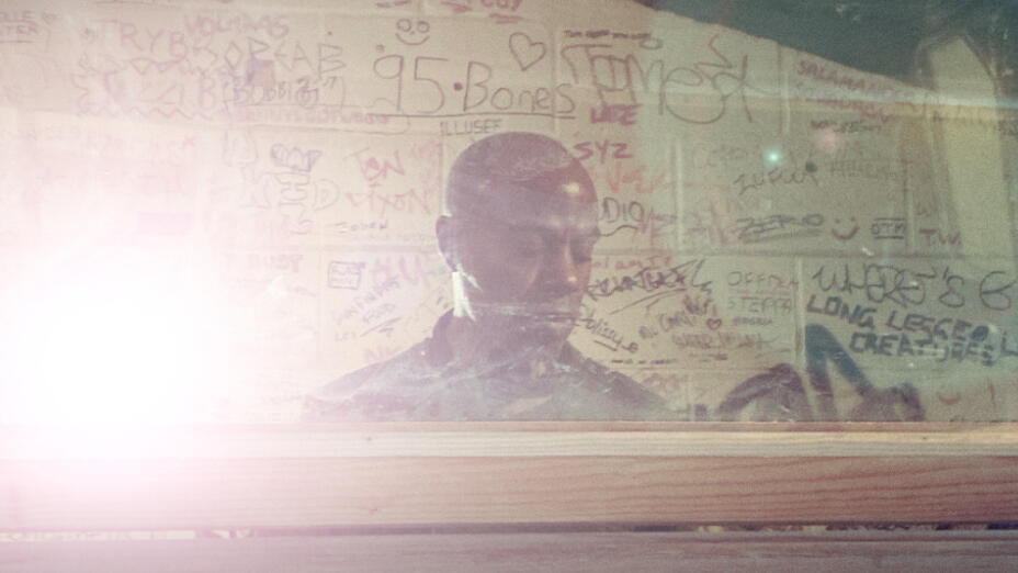 Composer Edward George sits behind a wooden desk in front of a white wall with graffiti.