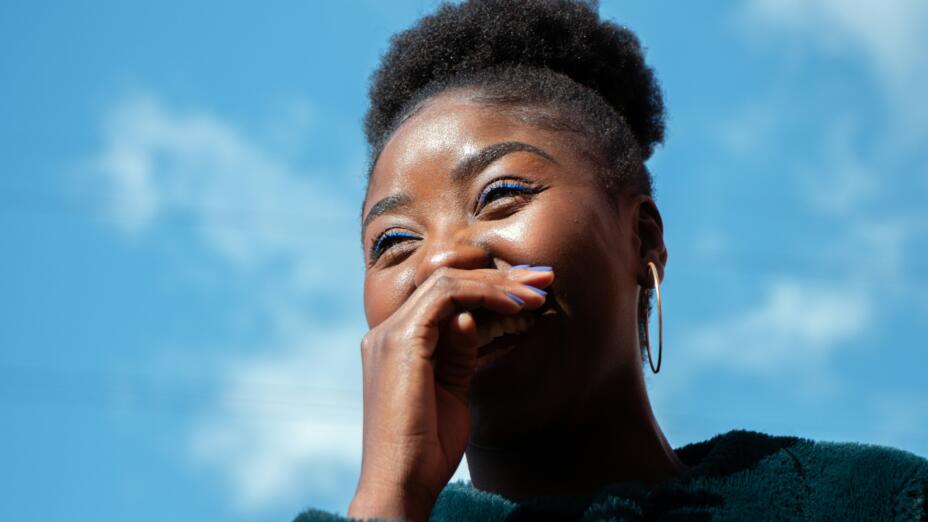 Young woman with hair up and gold earrings laughs and holds her hand over her mouth in front of a blue sky.
