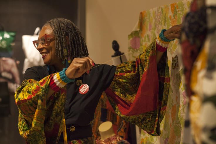 A smiling woman is holding string up to a fabric artwork. The artwork isn't in frame, but the woman is wearing brightly patterned dress with flowing fabric and a badge that says 'Big Ting'