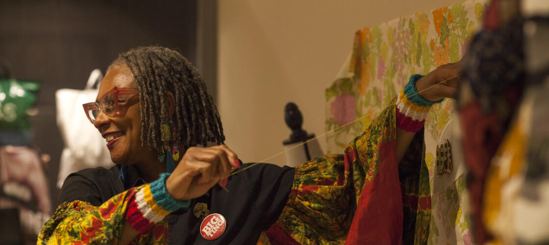 A smiling woman is holding string up to a fabric artwork. The artwork isn't in frame, but the woman is wearing brightly patterned dress with flowing fabric and a badge that says 'Big Ting' Photo credit: Elzbieta Piekacz