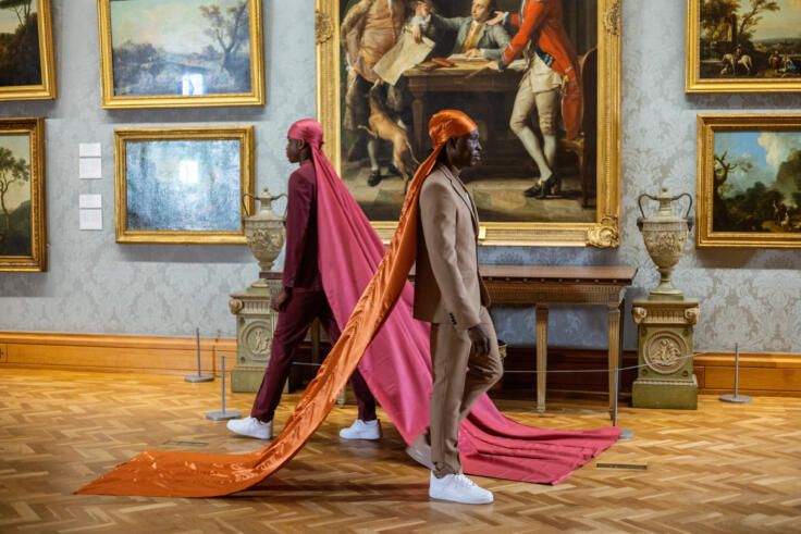 Two young black men are walking past each other. In the background, there are ornate 18th and 19th century paintings in gold frames from the National Museum Wales collection. The young man on the left is dressed in a plum red suit and a similar coloured durag which flows down into floor-length silk. The man on the right is in a tan suit, with a bright orange durag which similarly flows down into floor-length silk