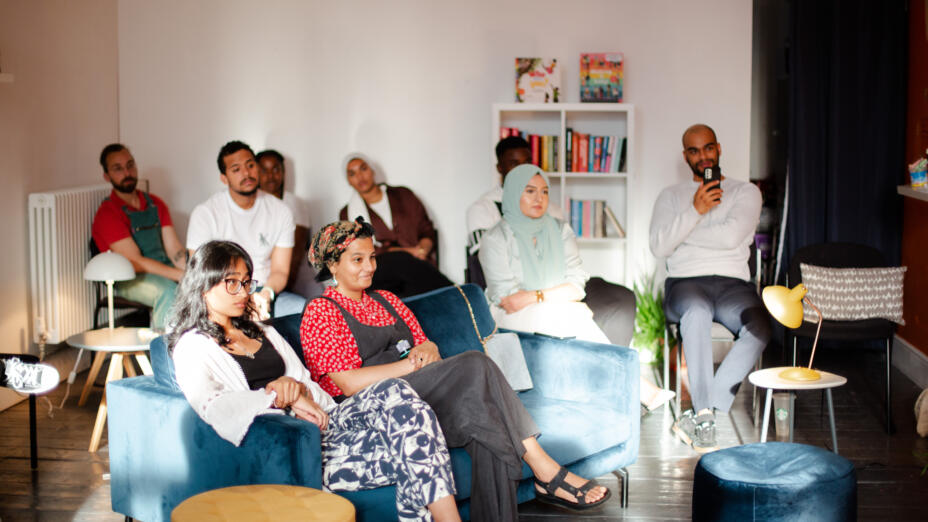 A group of nine people are sitting in a bright white room, on a range of cosy seating looking in the same direction and listening to a speaker who is out of frame.