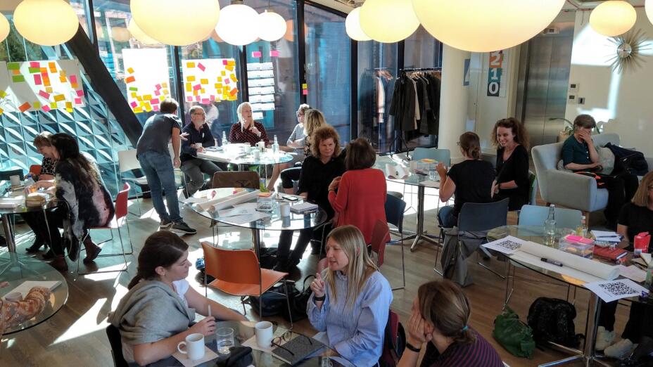 Group of teachers sitting in groups of 2 or 3 at small white round tables talking in an animated way. Large globe lamps hang from the ceiling and big pieces of flip chart paper are covered with notes on colourful post-its.