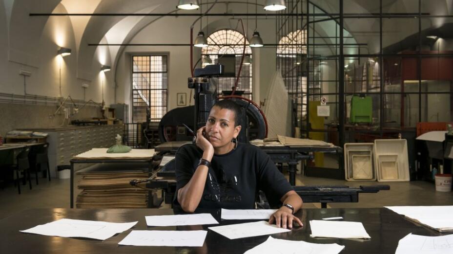 Artist Helen Cammock sits at a desk with papers in front in a library.