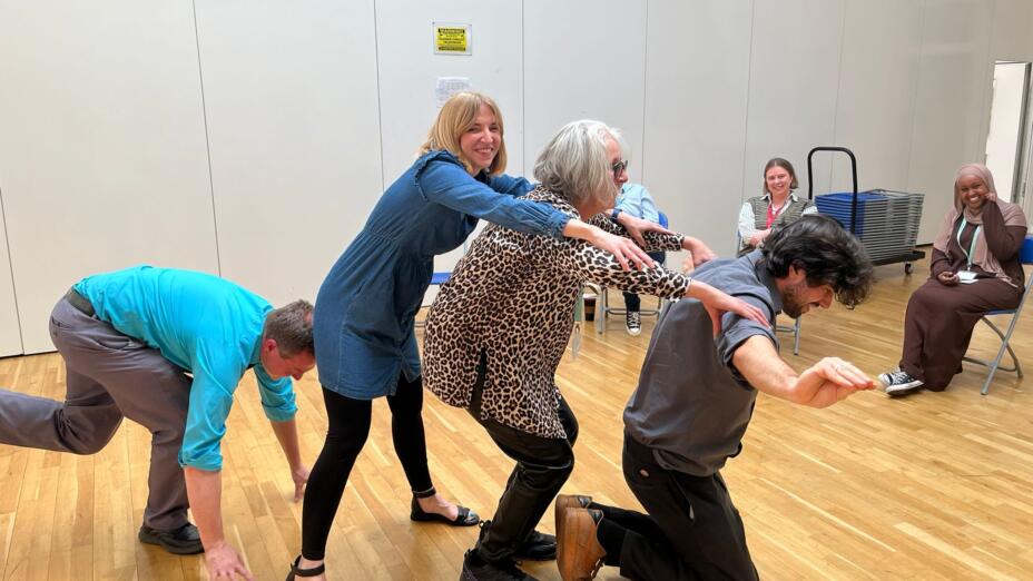 Six teachers take part in an arts-based continuing professional development session in a school assembly hall with a wooden floor.
