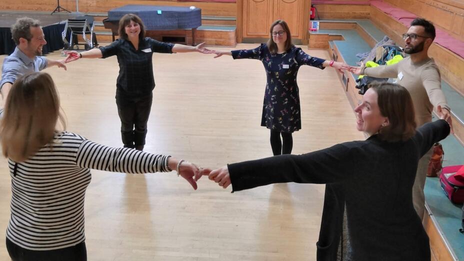Group of six teachers stand in a wide circle holding hands happily in a wood panelled assembly hall.
