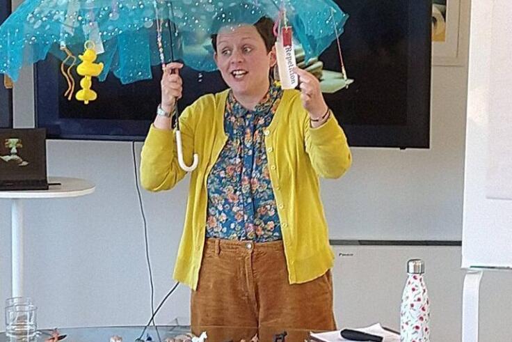 Young woman in a yellow cardigan stands in front of a screen on a wall. She is holding a turquoise translucent umbrella and standing by a glass table.