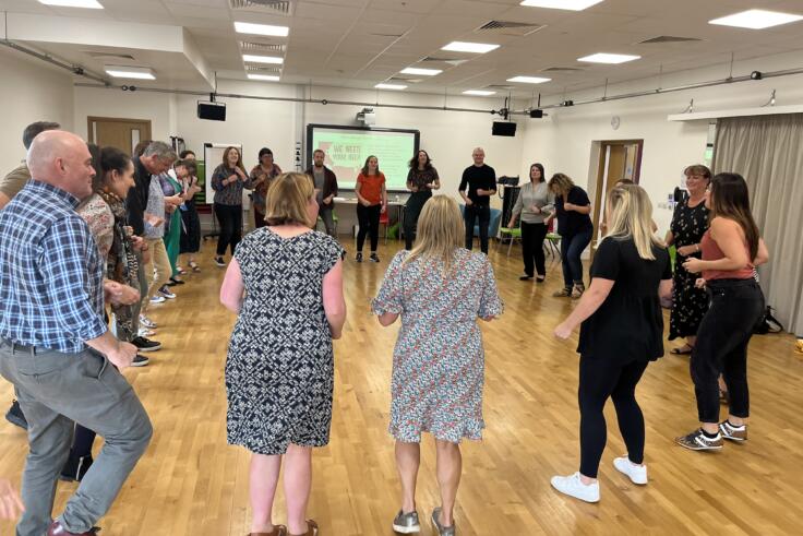 Big group of teachers standing in a circle in a room with wood floors as they take part in a drama-based CPD session.