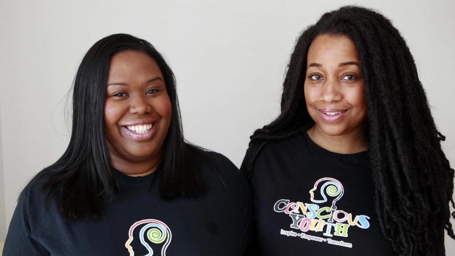 Two young women smiling to the camera, wearing Conscious Youth t-shirts that say inspire, empower, transform.