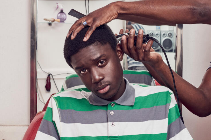 Young man sits in a barber chair as he gets his hair cut with clippers. He's wearing a green, white and grey polo shirt.