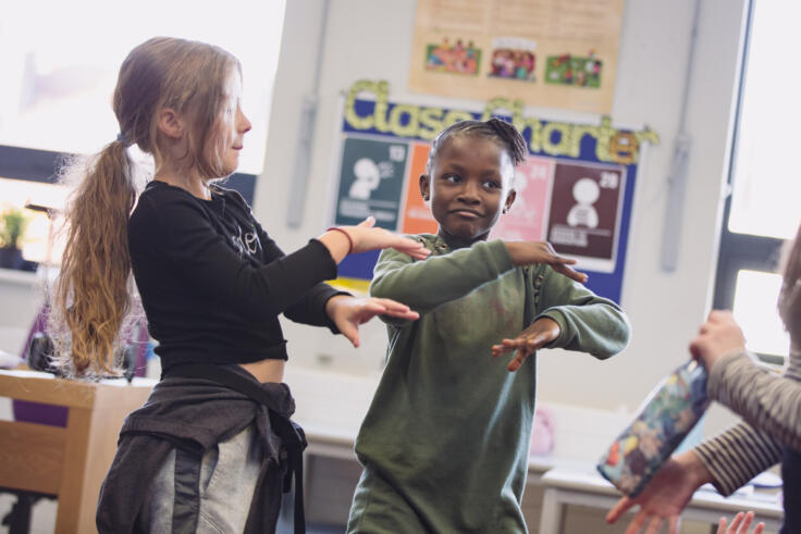Two young girls are in a classroom, mid-dance. They have their arms crossed out in front of them, one above the other
