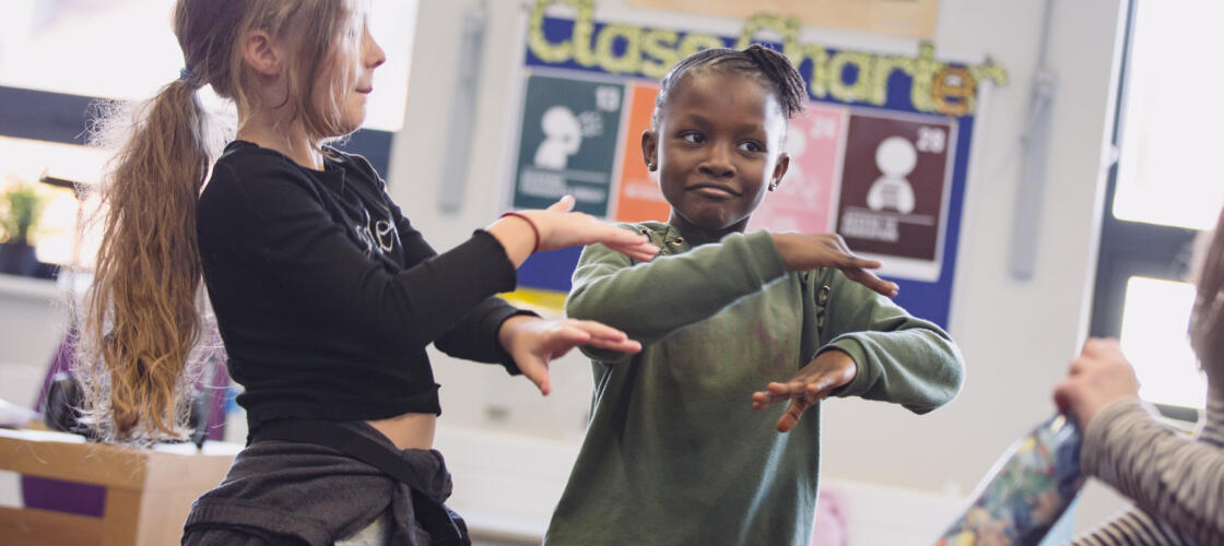 Two young girls are in a classroom, mid-dance. They have their arms crossed out in front of them, one above the other Photo credit: Camilla Adams