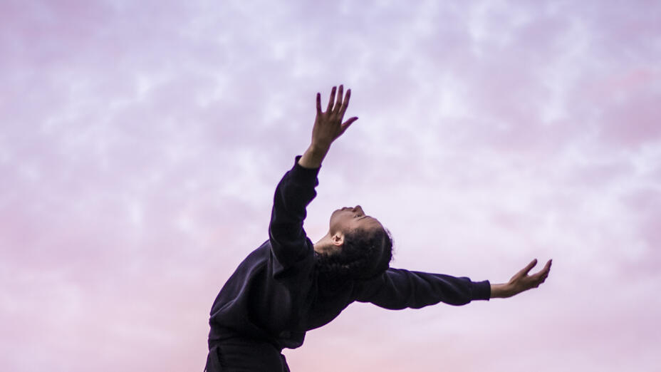 A dancer is posing against a bright but cloudy sky. They are slightly silhouetted, facing away from the camera with their head tilted back and arms reaching out behind them